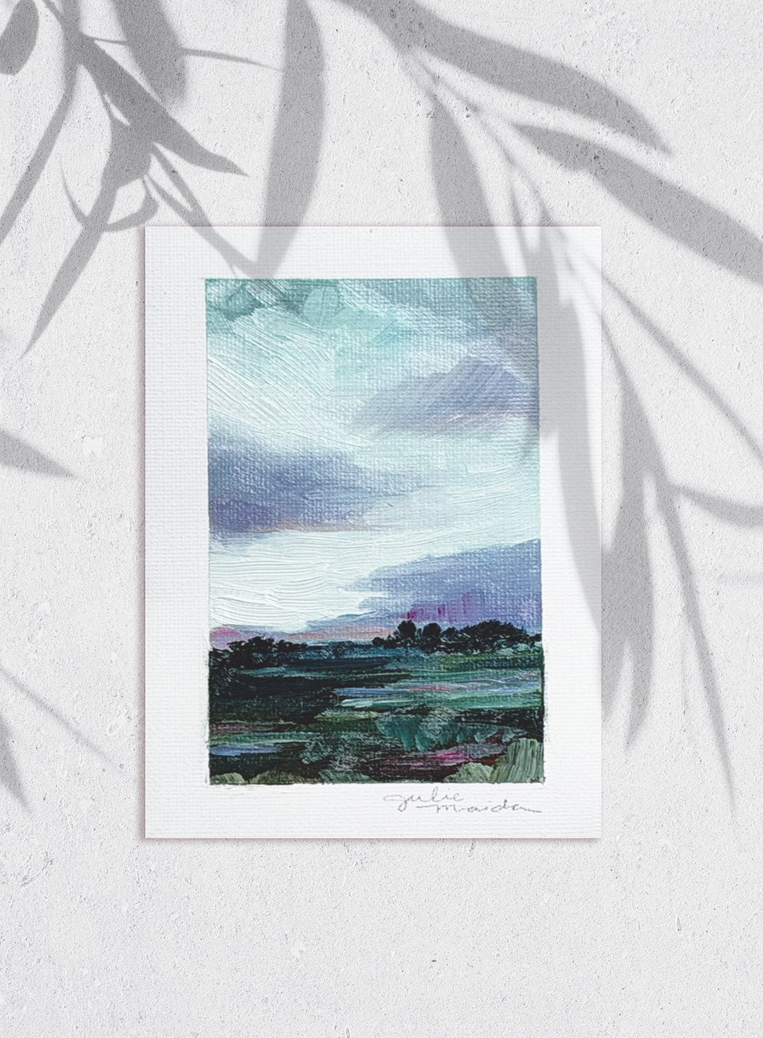 A vertical art print featuring a textured landscape painting with a blue and purple sky, green fields, and dark tree silhouettes, displayed on a light textured wall with leaf shadows cast across it.